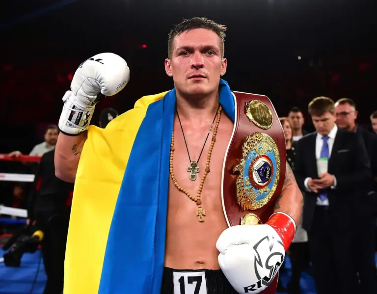Oleksandr Usyk wearing boxing gloves and a championship belt, draped in the Ukrainian flag.